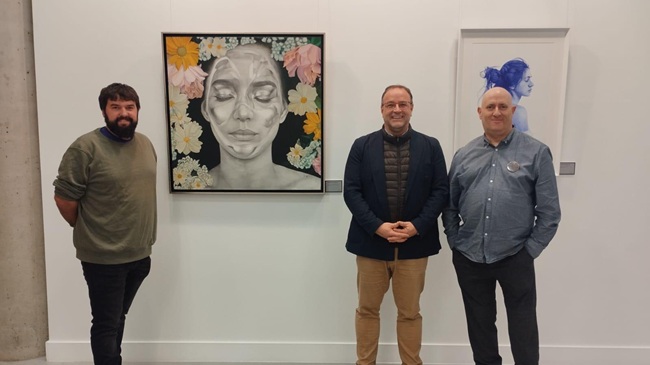 Tres hombres posan en la sala de exposiciones del CMI El Charro junto a dos cuadros de la muestra ‘Ventanas a la emoción II’ de David Sánchez. A la izquierda, un retrato realista rodeado de flores; a la derecha, un perfil femenino en tonos azules.