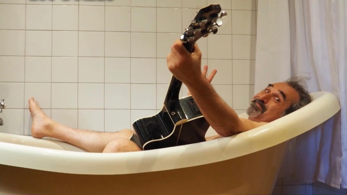 aniel Higiénico posando de forma cómica en una bañera con una guitarra acústica, representando su estilo irreverente para el show en Oviedo.
